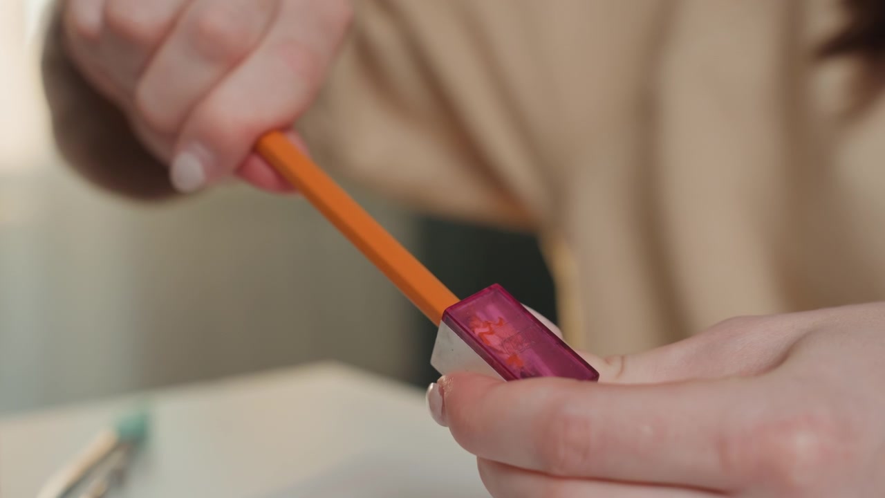 Manos caucásicas afilando un lápiz en el escritorio de casa, tutora preparando materiales de práctica de gramática, detalle cercano de un lápiz naranja y un sacapuntas morado, virutas limpias cayendo sobre la mesa blanca, luz natural cálida.