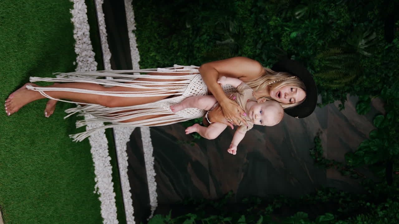 Woman in black hat and white knitted suit holding her little infant child. Mom plays and tosses her baby. Vertical screen.