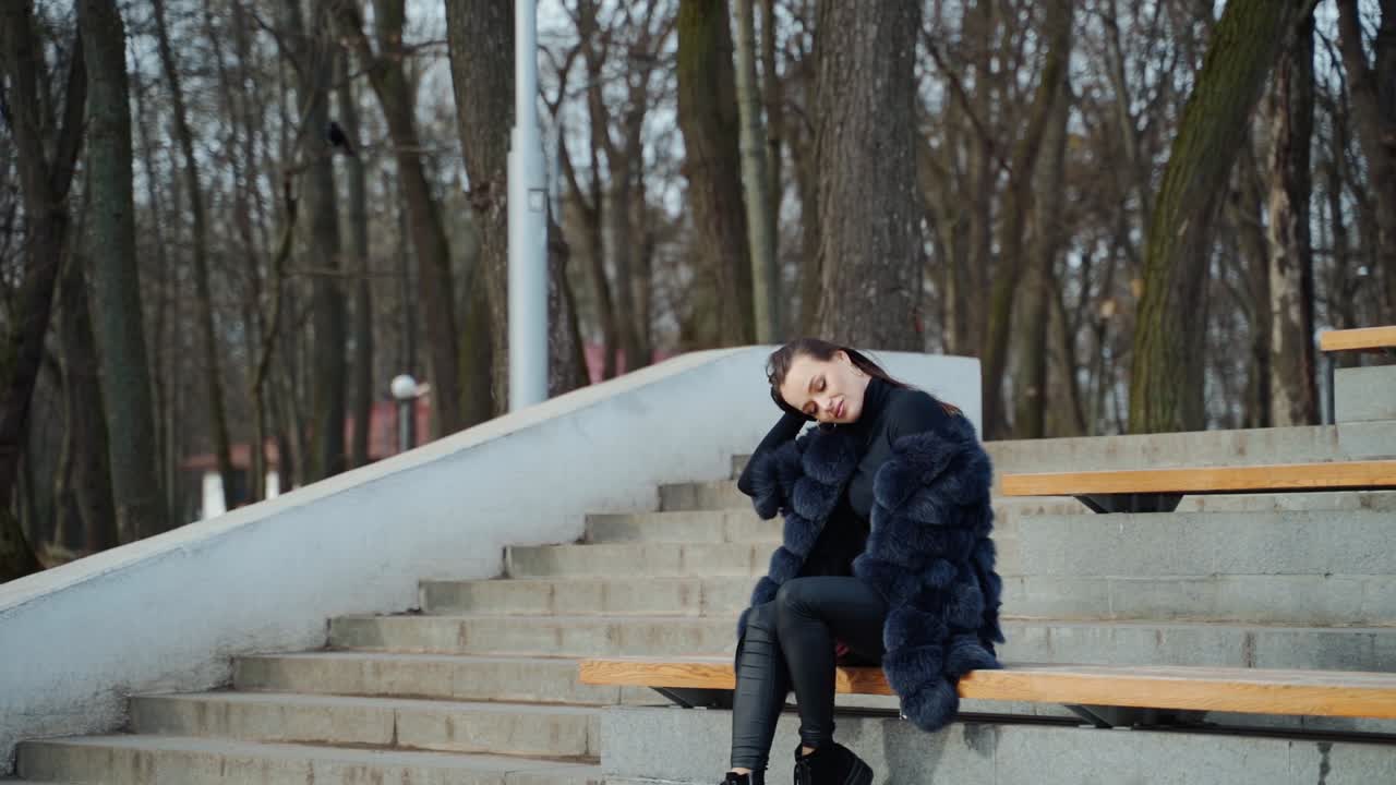 Beautiful woman sitting outdoors on the background of leafless trees. Side view of a sexy woman in black fur coat resting in the park. Slow motion.