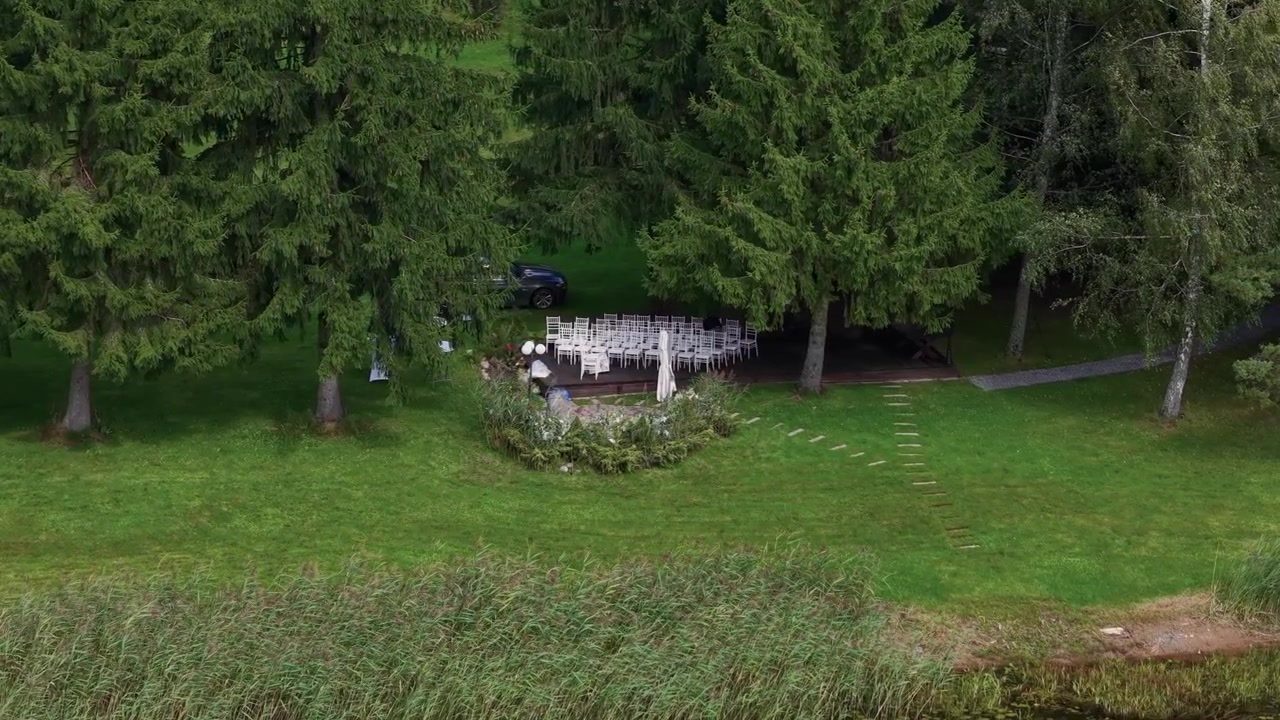 Aerial footage captures an empty wedding ceremony setup with white chairs and decor nestled by tall trees near a serene lake, under soft daylight and calm, natural surroundings.
