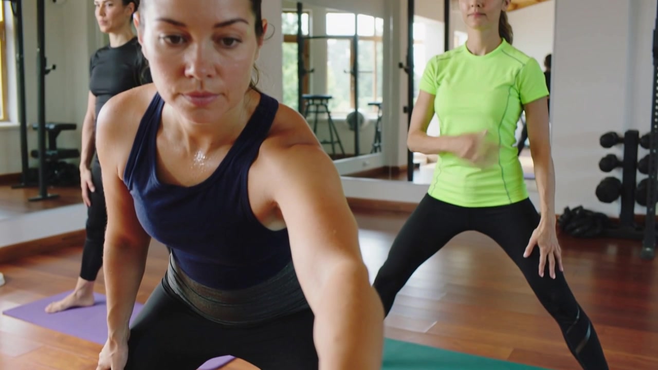 Mujer con top azul marino extendiendo la mano para presionar su reloj deportivo sobre una colchoneta doblada en un estudio de fitness, comprobando la hora de la clase.