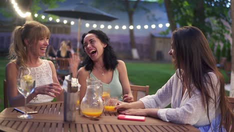 three female friends sit in the outdoors cafe, drink juice and have fun communicating, laughing in the evening