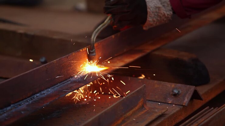 In primo piano, processo di saldatura industriale con scintille luminose, metallo incandescente e travi d'acciaio. Lavoro manuale nell'officina di fabbrica. Concetto di industria pesante, produzione, costruzione e riparazione.