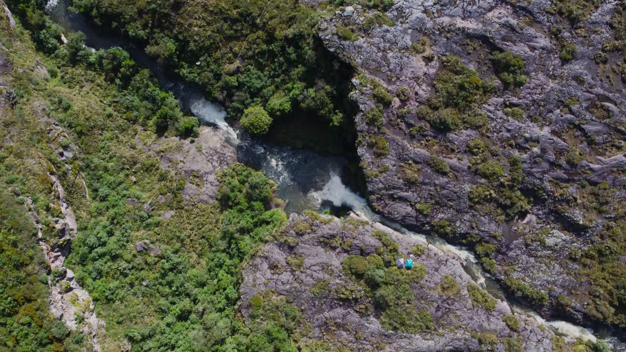 A breathtaking aerial top-down dolly-in shot approaching Fuego Waterfall near Cotopaxi Volcano, Ecuador. A lone tent is pitched beside the rushing waters, showcasing an isolated campsite in nature.