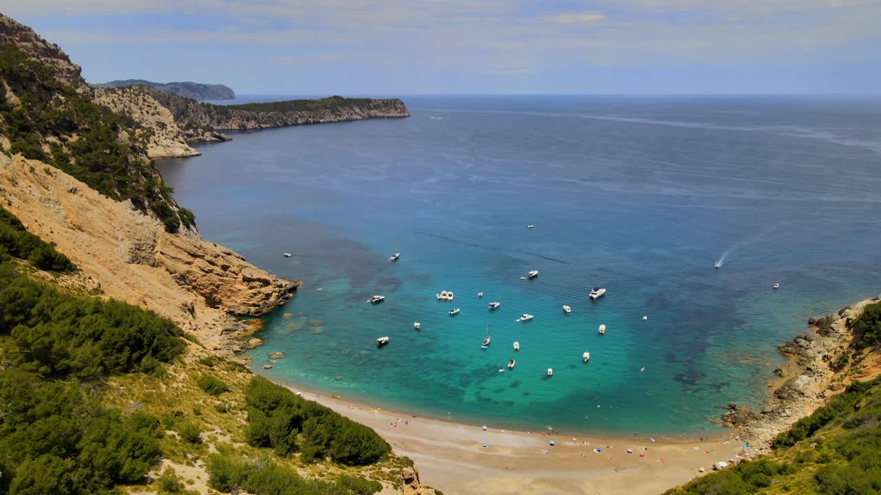 Enjoy the stunning coastal scenery of Coll Baix in Puerto de Alcudia, Majorca. The clear blue waters are dotted with boats, surrounded by beautiful cliffs and lush greenery.