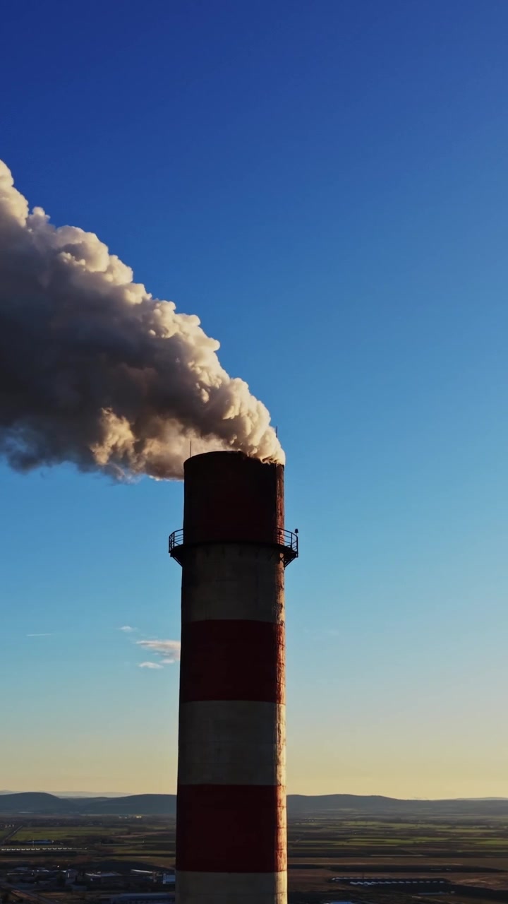 Fumo che si alza da un'alta ciminiera di fabbrica al tramonto sopra una zona industriale