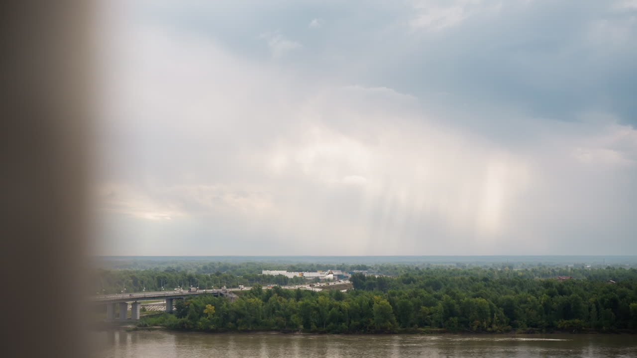 비구름 아래 강 풍경, 멀리 보이는 녹색 수목선과 강 위로 쏟아지는 빗줄기, 왼쪽에 콘크리트 다리, 잔물결이 있는 반사되는 물 표면, 부드러운 햇살이 비치는 흐린 하늘, 영화 같은 분위기
