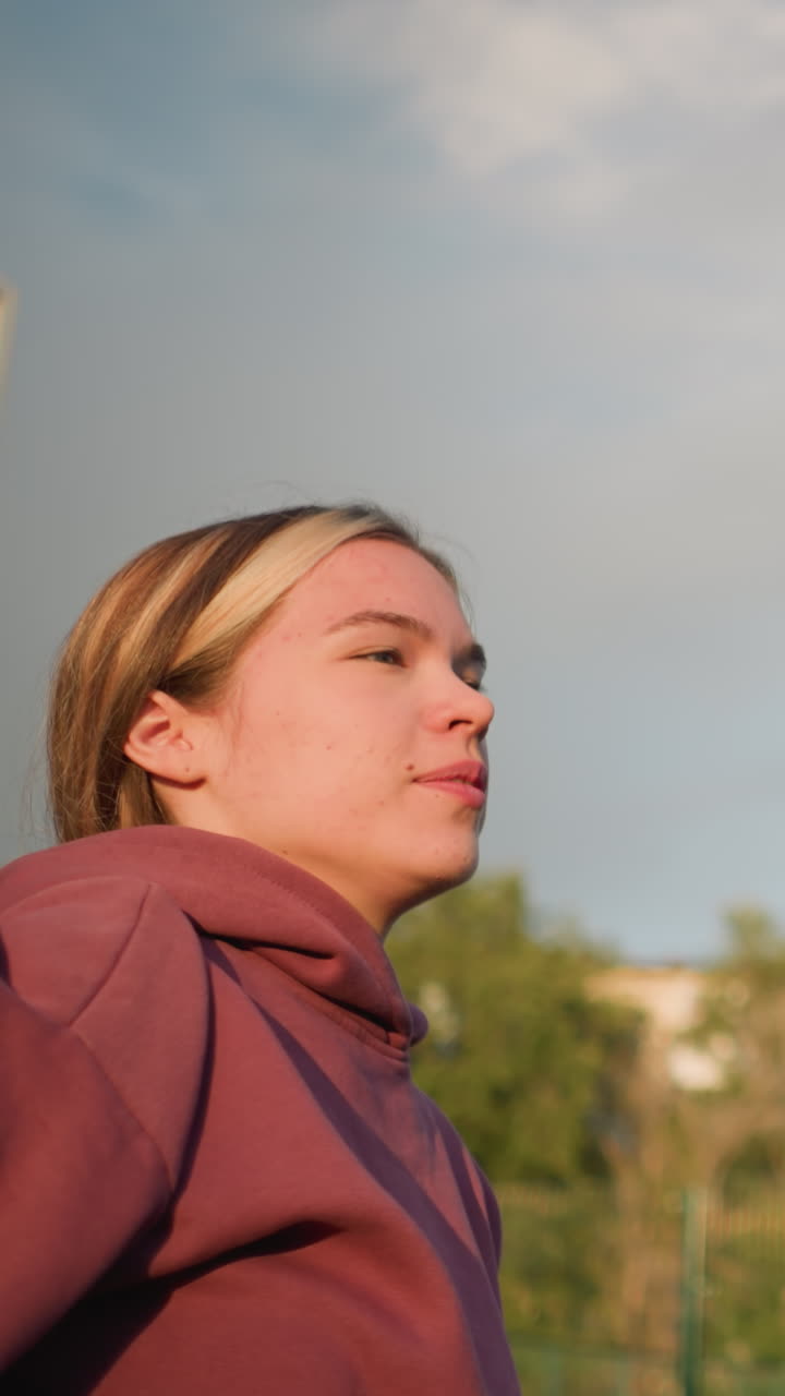 Giovane donna con felpa che serve a pallavolo all'aperto con canestro da basket visibile sullo sfondo, che si prepara a servire in un campo aperto con un'espressione calma e concentrata e luce naturale