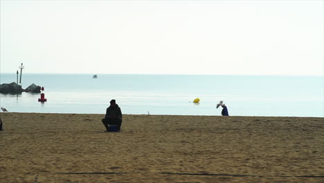 silueta en cámara lenta de la gente en la playa de la barceloneta, sentada y caminando, con pájaros volando junto a un muelle rocoso