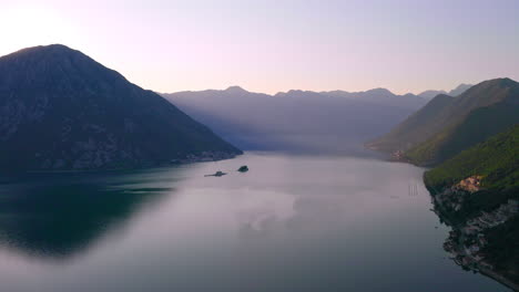The-sun-slowly-rising-from-behind-a-high-mountain-above-the-still-waters-of-Kotor-Bay,-Montenegro,-surrounded-by-hills-and-mountains,light-morning-haze-above-the-whole-area,cloudless-sky,-aerial-shot