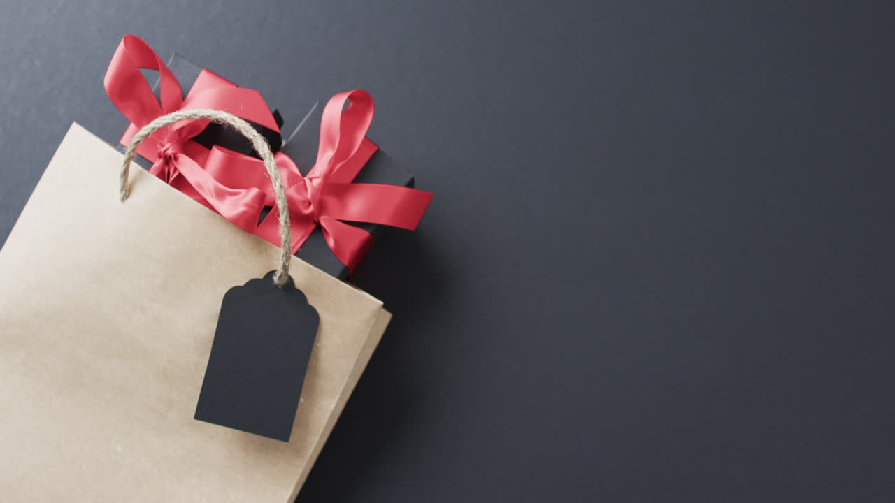 una borsa regalo di carta marrone con un nastro rosso vibrante e un tag vuoto si trova su una superficie scura, con copia s
