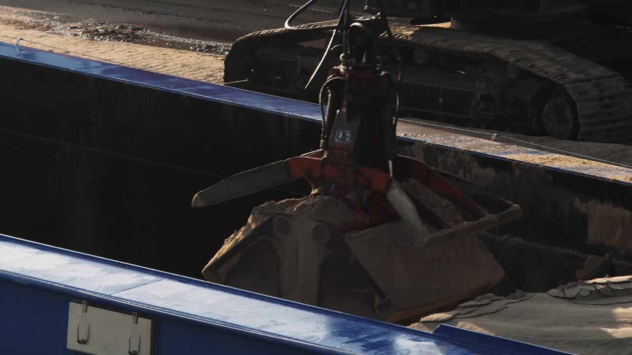 An excavator with a clamshell bucket unloads sand from a cargo barge at an industrial port on a sunny day