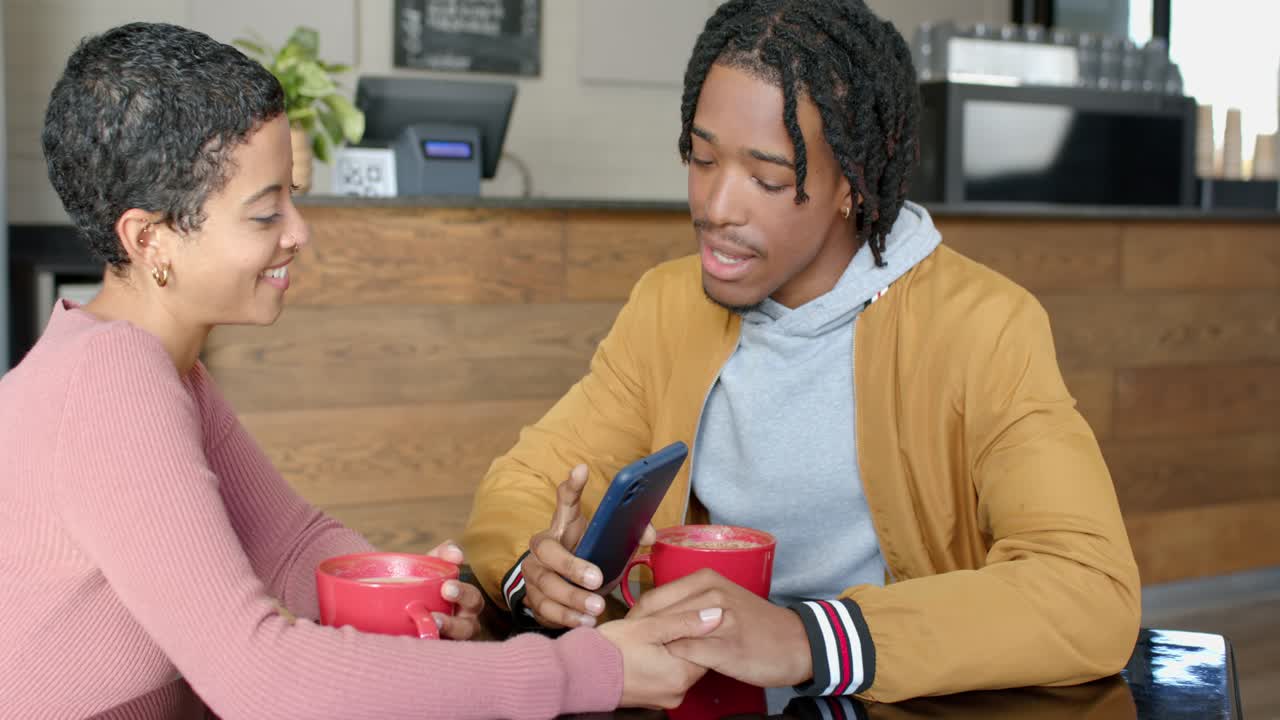 Diverse couple clasping hands at cafe table, lifting smartphone and sharing screen beside red mugs