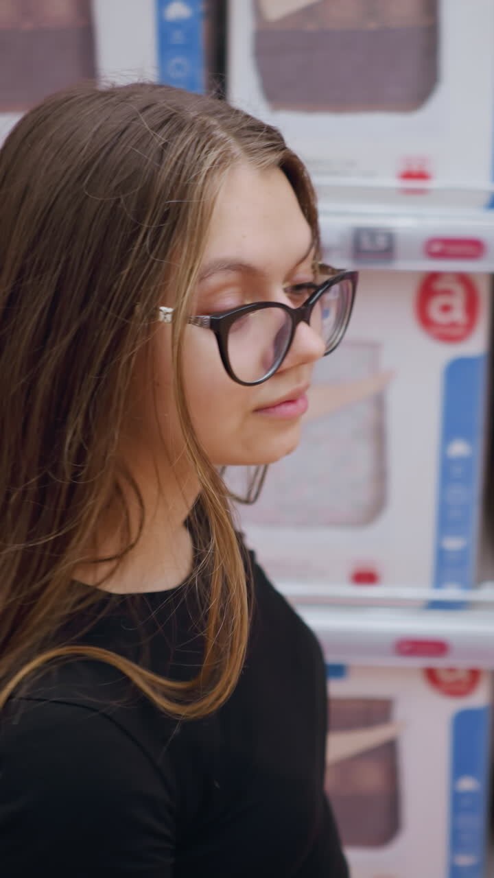 Aerial close-up of young woman walking by a store shelf filled with bedding products, she observes different designs, evaluating comfort and style options, retail shopping experience
