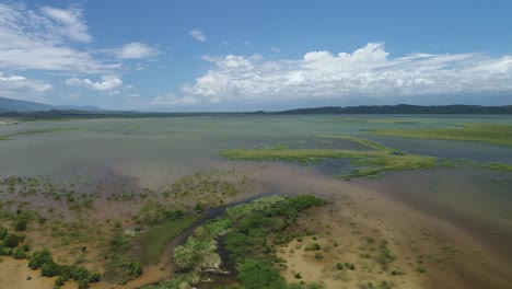 Sandy-grass-and-shrubs-grow-into-the-ocean-waters-surrounded-by-mountains-on-a-sunny-day,-drone-aerial