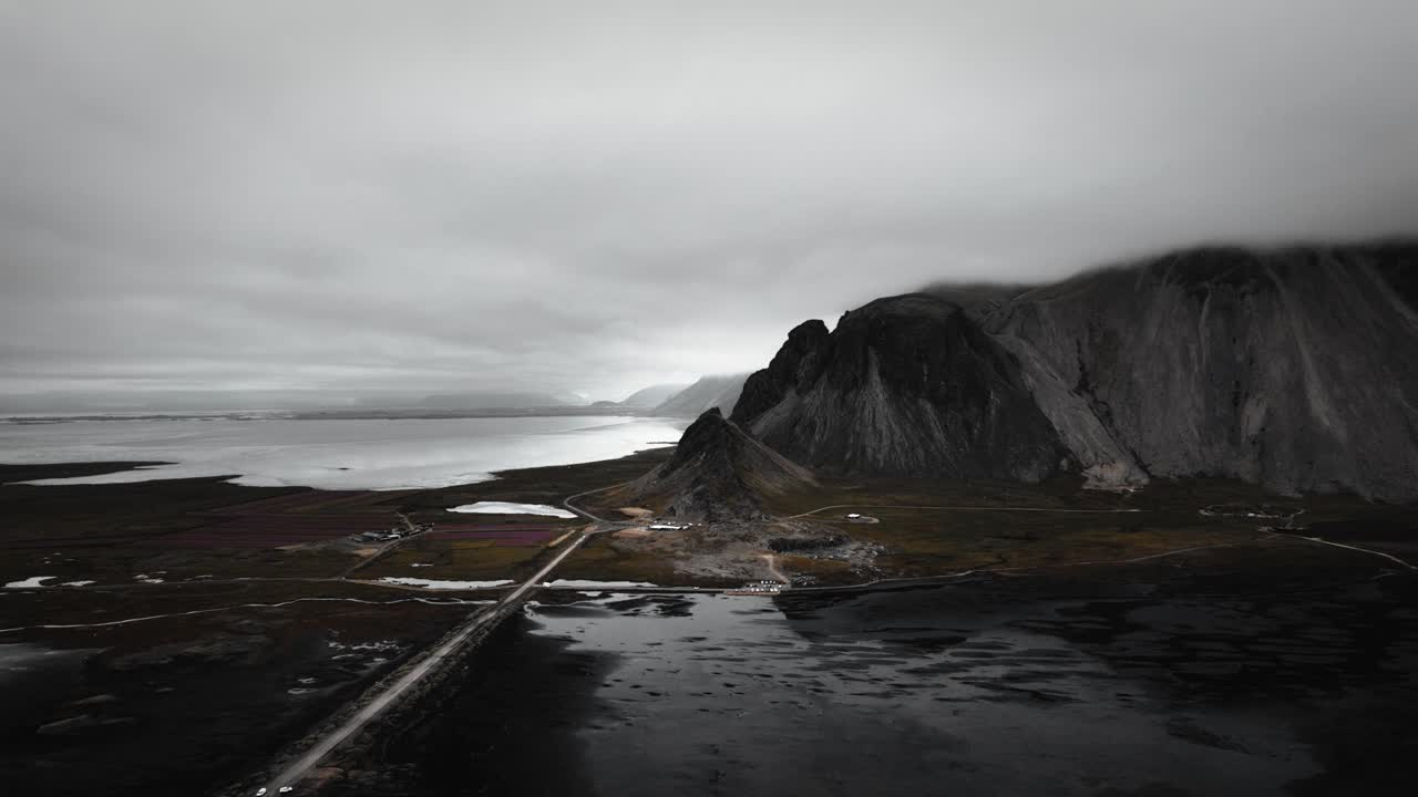 vuelo aéreo sobre playa de arena negra stokksnes, montañas volcánicas oscuras y carretera en la distancia, paisaje oscuro de mal humor nublado islandesa