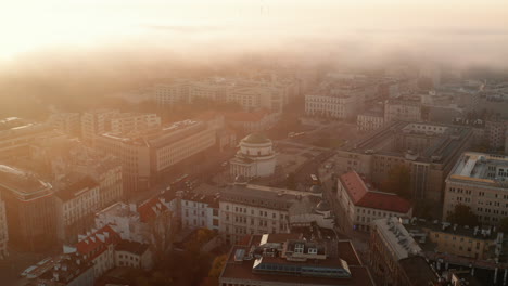 Aerial-view-of-buildings-in-town-in-foggy-morning.-Bright-golden-rising-sun.-Church-on-Three-Crosses-Square.-Warsaw,-Poland