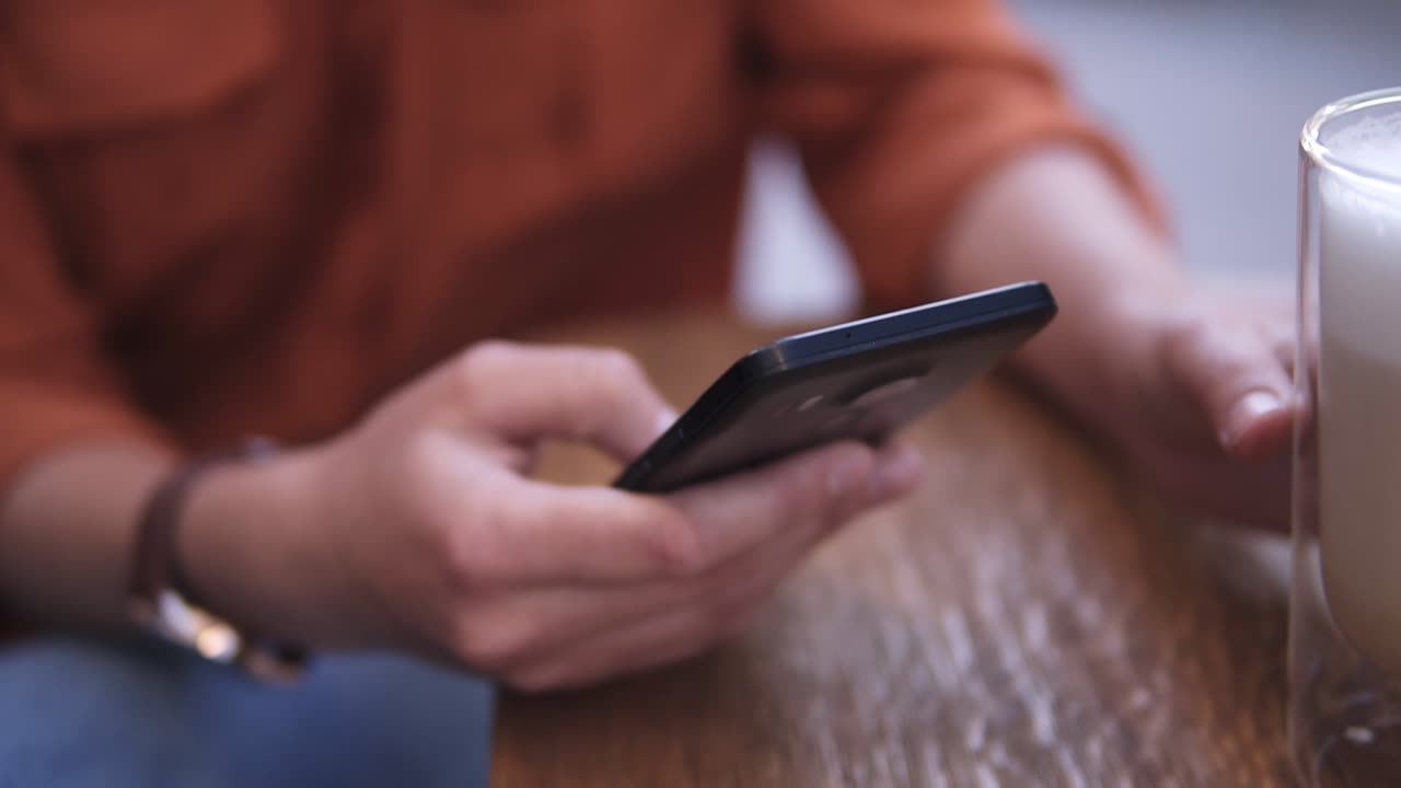hermosa joven rubia con gafas elegantes sentada en un café al aire libre, bebiendo capuchino y desplazando su teléfono móvil