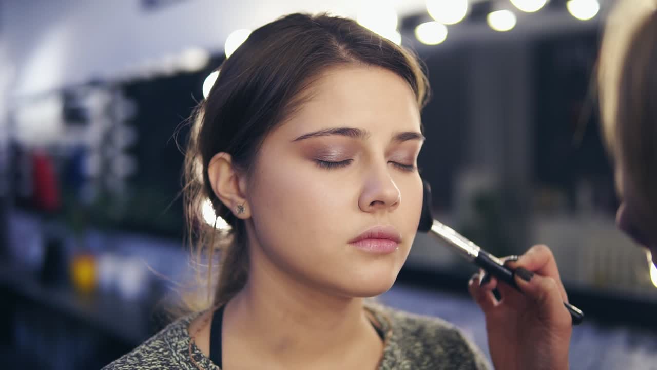 Close Up view of professional makeup artist's hands applying facial powder on young woman's skin using special brush. Slow Motion
