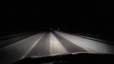 headlights cut through the dark as fine snow drifts down, on a lonely snow-covered road under the night sky