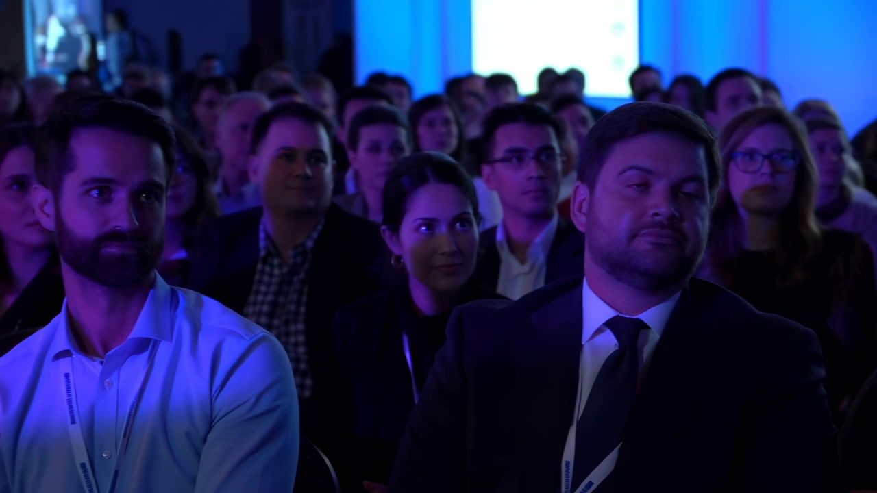 Narrativa cronológica de la audiencia a lo largo del clip: la apertura muestra filas de profesionales sentados bajo una iluminación de escenario azul fría, con la presentación en curso.