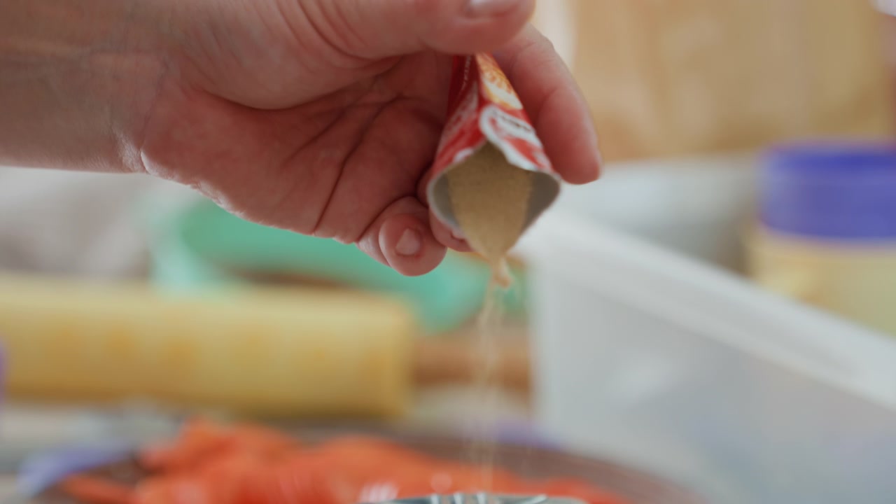 Nahaufnahme einer Küchenszene aus niedrigem Winkel, die eine Hand zeigt, die eine kleine Gewürzpackung aufdrückt, während ein dünner Saucenstrahl und Tropfen in einen Topf oder eine Schüssel fließen, mit sichtbarer Verformung der Packung und Kochbewegung.