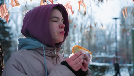 vista lateral de una mujer comiendo maíz con hojas secas balanceándose suavemente por encima, fondo borroso con personas caminando en el parque durante el invierno, luz natural suave reflejándose en su cara, momento pacífico del parque
