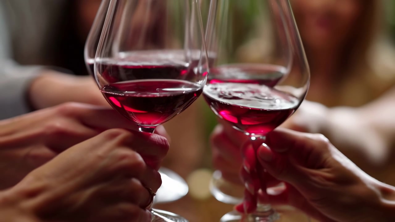 Primer plano de varias manos levantando y tocando copas de vino llenas de vino tinto, con un fondo suave y cálido que sugiere una reunión festiva en interiores