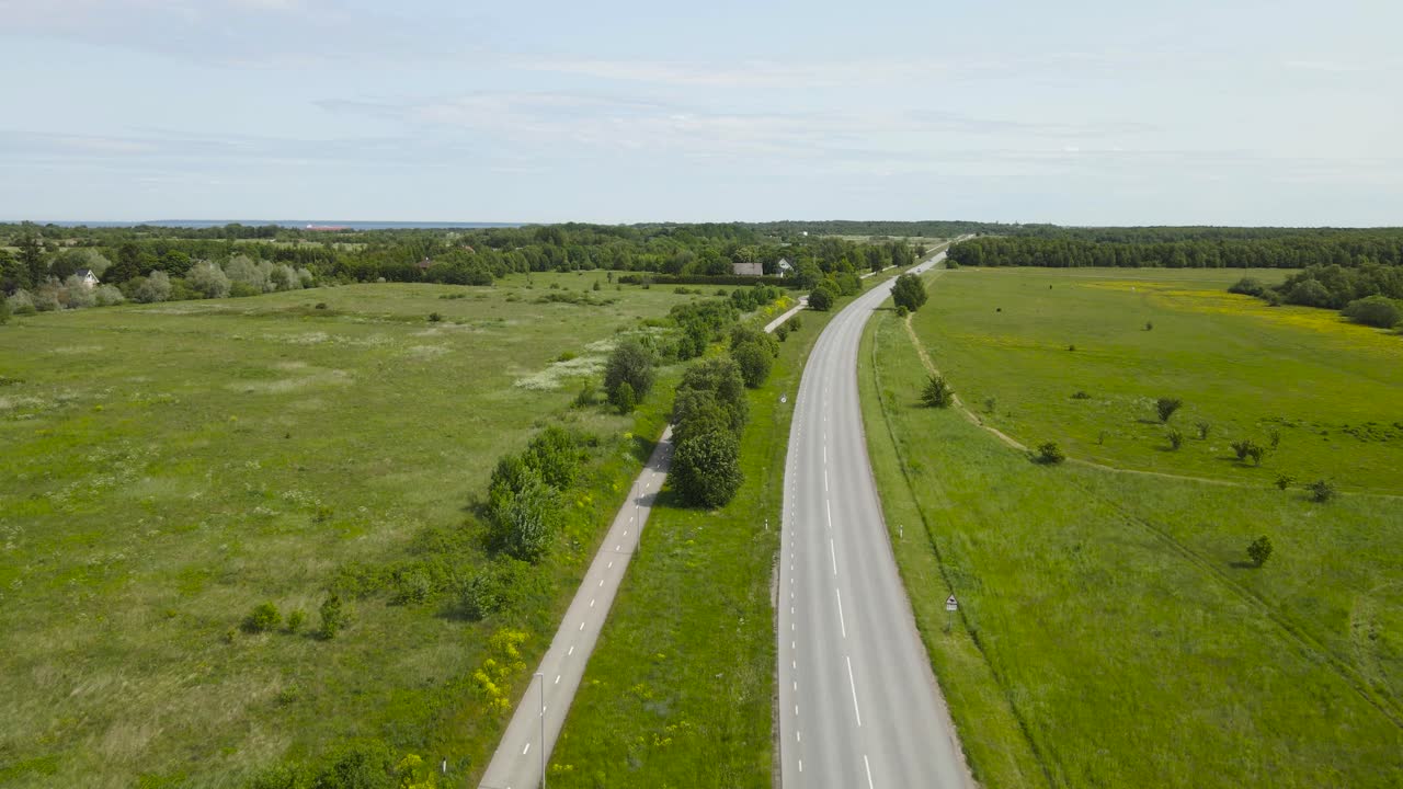 Aerial drone footage flying over a rural countryside road where cars, vehicles and trucks can be seen driving during summer sunny day. Bicycle and pedestrian road on the side separated with grass area