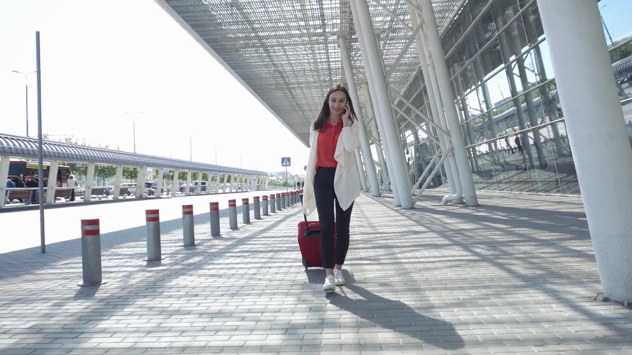 Mujer viajera sonriente tirando de una maleta y hablando por smartphone fuera del aeropuerto