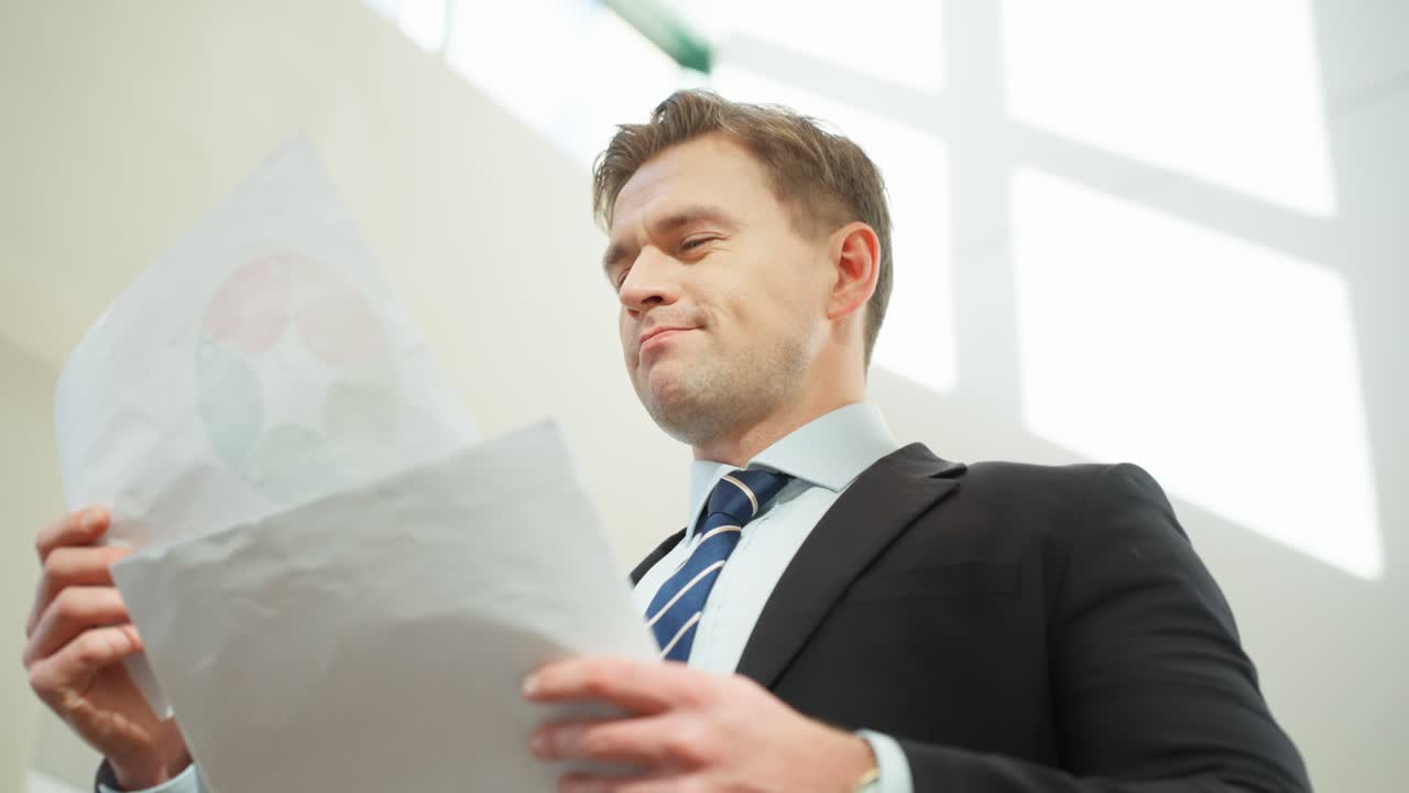 Successful manager man holding and examining paper sheet documents in a brightly lit office environment - low angle close-up
