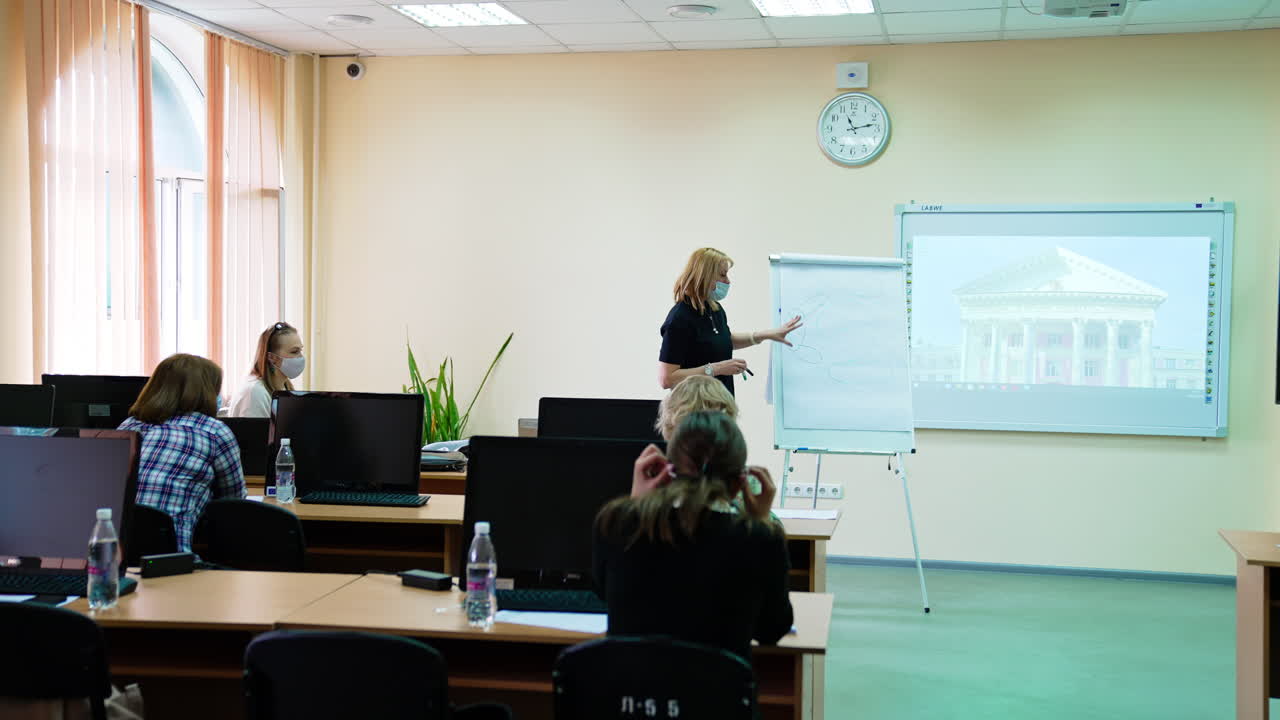 Female lecturer speaking, explaining and pointing at the blackboard. Giving a lecture to the auditory of students.