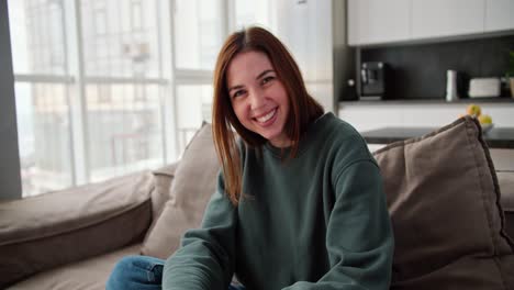 Portrait-of-a-happy-brunette-girl-smiling-widely-in-a-green-sweater-and-jeans-sitting-on-a-modern-brown-sofa-in-the-apartment