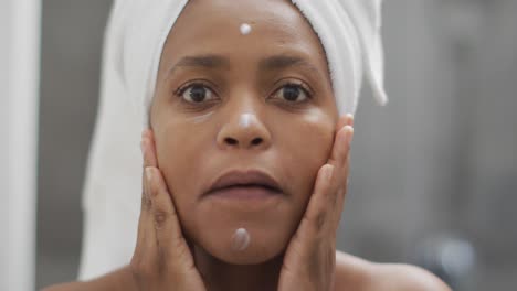 Happy-african-american-woman-apply-cream-on-face-in-bathroom