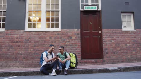 Two-mixed-race-male-friends-sitting,-talking-and-using-smartphone-in-the-street