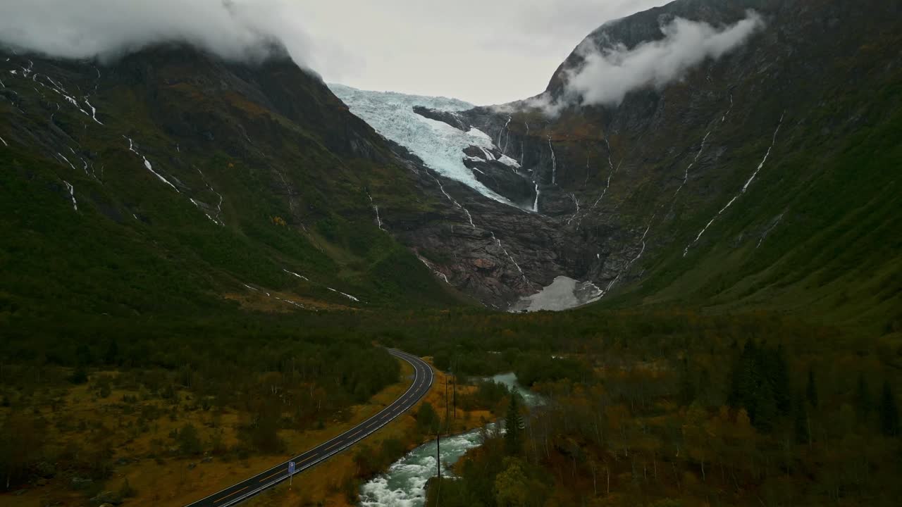 drone vuela a través de un valle sobre una carretera noruega hacia un glaciar cubierto de nubes