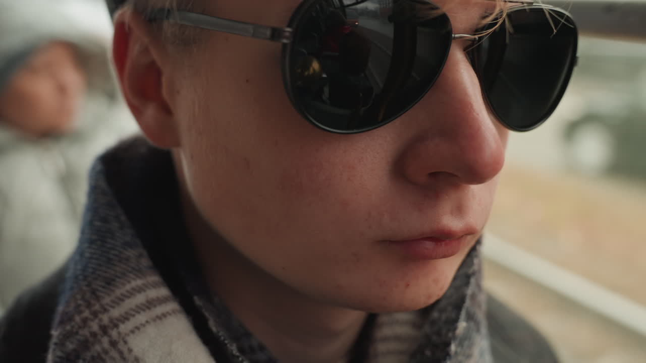Primo piano di un giovane creativo bianco con occhiali da sole, sciarpa, al finestrino di un autobus, lenti riflettenti e sfondo cittadino dai colori tenui. Regista indipendente in viaggio, espressione contemplativa, toni neutri cinematografici, sottile