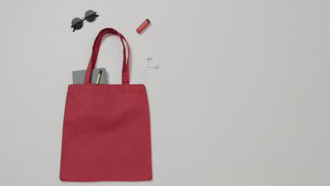 bolsa roja con gafas de sol, portátil, lápiz labial, auriculares sobre fondo blanco, espacio para copiar, cámara lenta