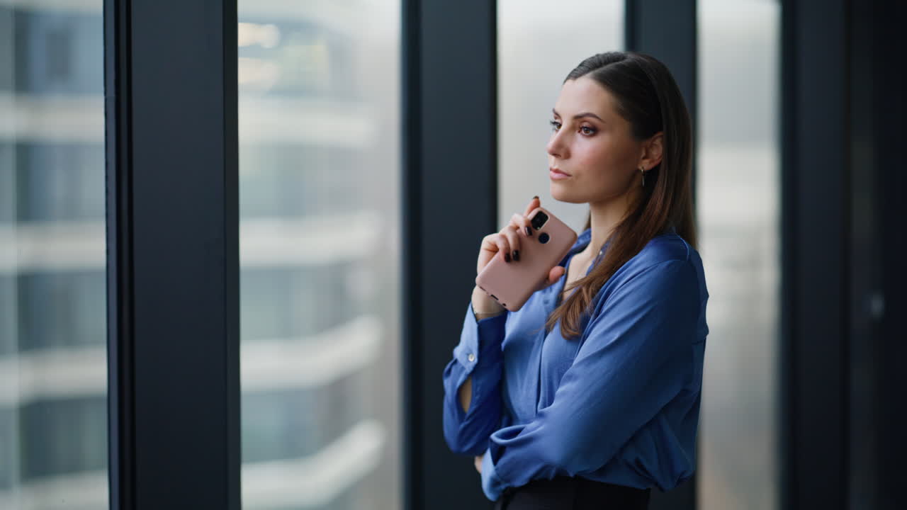 Thoughtful business owner standing by office window holding smartphone closeup