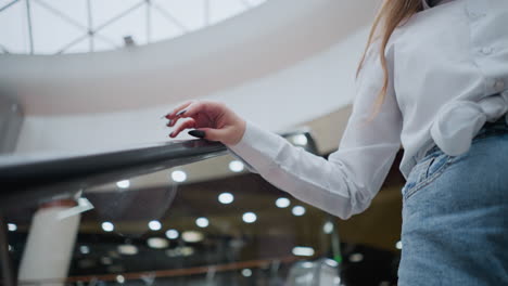 woman with black polished nails gently taps escalator handrail while descending, she wears a white blouse and jeans, soft-focus background