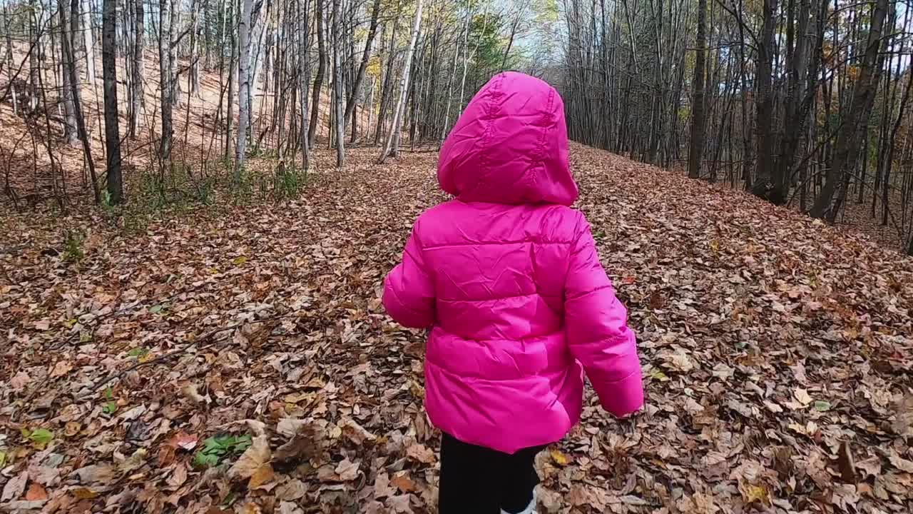 siguiendo a una niña pequeña con chaqueta rosa con capucha mientras corre por un sendero cubierto de hojas en el bosque y las hojas vuelan a cámara lenta