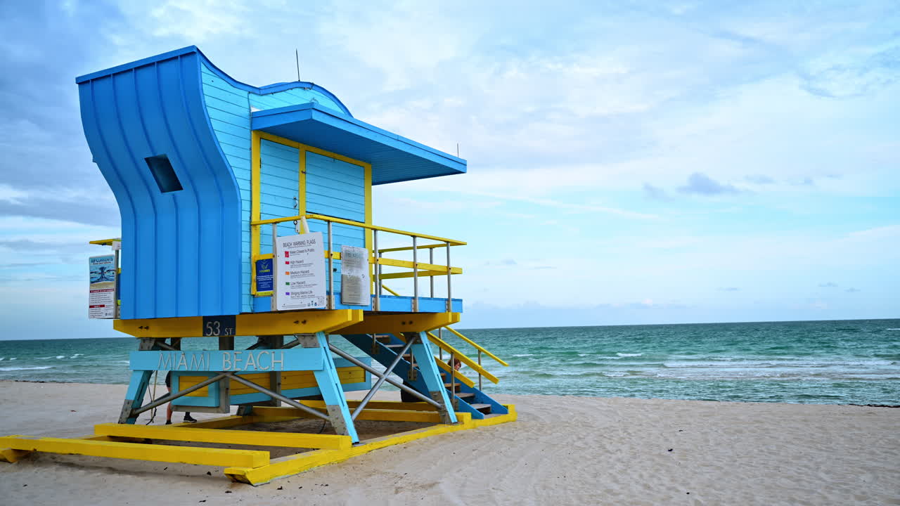 Torre di salvataggio blu e gialla sulla sabbia di Miami Beach. Iconica stazione di soccorso in legno sulla battigia sotto un cielo nuvoloso con onde oceaniche sullo sfondo.