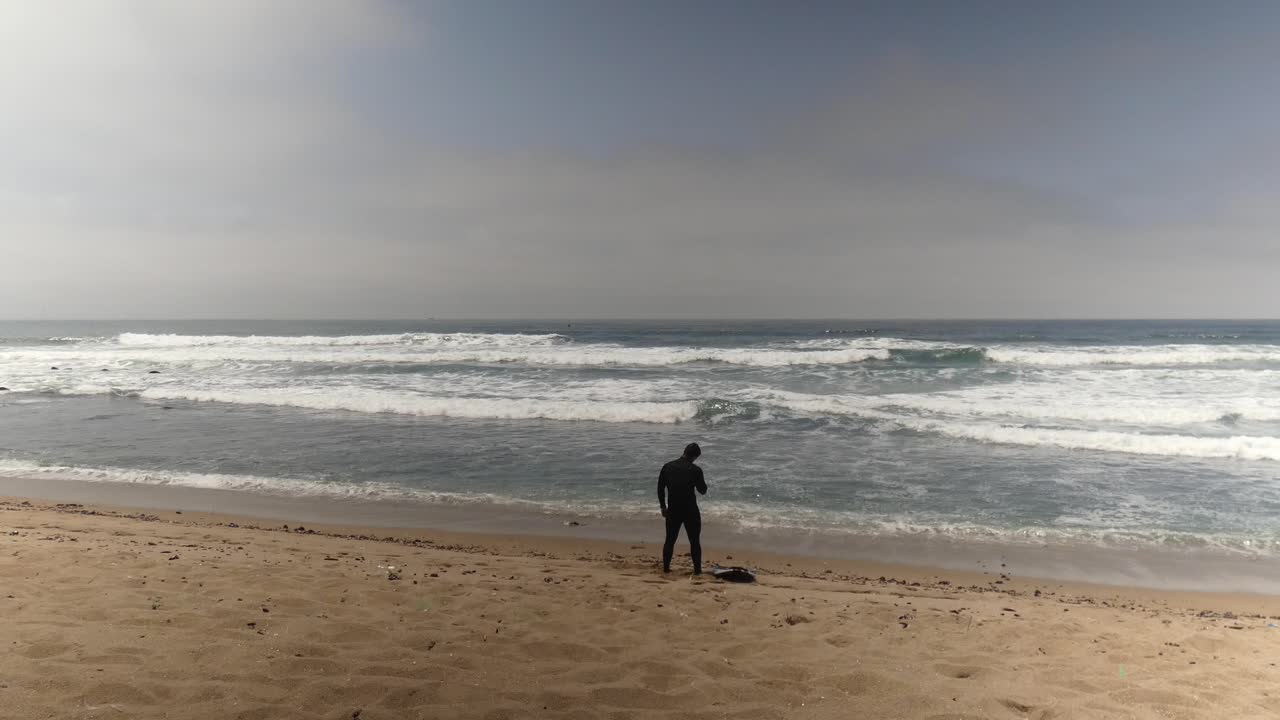 un surfista en la orilla de la playa de arena, estirándose, antes de recoger su tabla de surf y dirigirse al océano