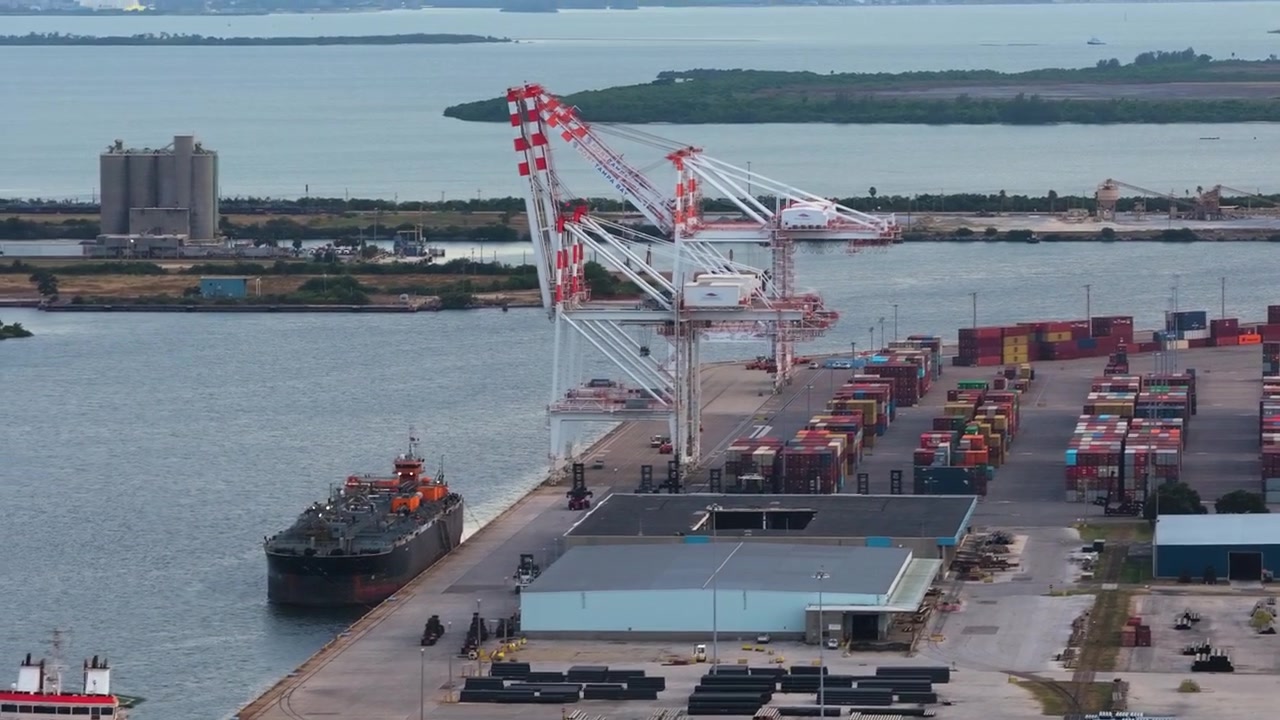 Port of Tampa container terminal with cargo stacks, cranes and docked ship, aerial orbit upward tilt
