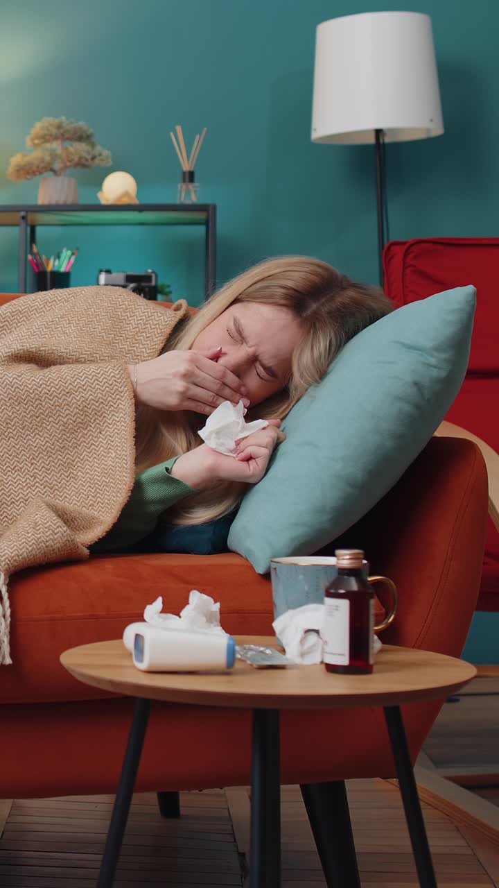 Sick ill young woman suffering from cold allergy lying on home sofa sneezes wipes snot into napkin