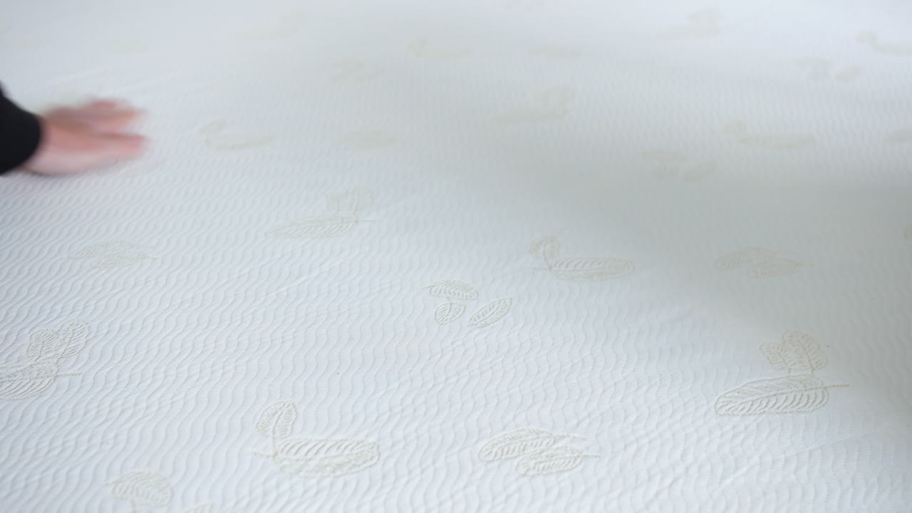 A hand pressing gently on a white mattress featuring embossed leaf patterns and subtle textured fabric. Close-up highlighting surface softness and fabric responsiveness to touch