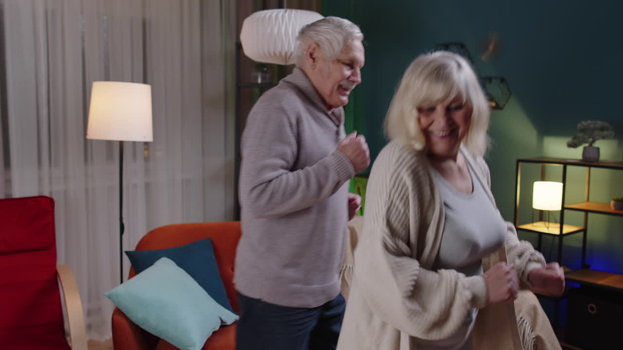 걱정 없는 노인 커플이 집에서 춤을 추고 휴식을 취하고 현대 음악으로 재미를 느끼고 집에서 웃고 있습니다.