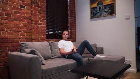 un hombre viene en el sofá, enciende la televisión con un control remoto y ve la televisión, y se relaja en la sala de estar en casa con una pared de ladrillo rojo