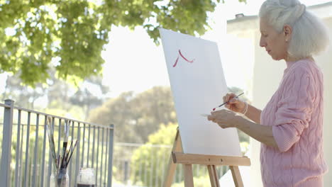 happy senior biracial woman painting at balcony at home, slow motion