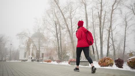 athlete walking along interlocked pathway with bag over shoulder, left hand touching chest while surrounded by snow-dusted benches, trees, light poles, and distant building in serene foggy atmosphere